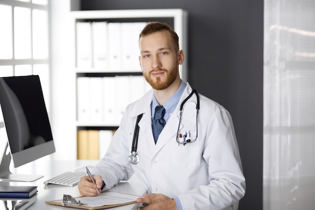 Friendly red-bearded doctor sitting and writing at clipboard in clinic. Perfect medical service in hospital. Medicine, healthcare.