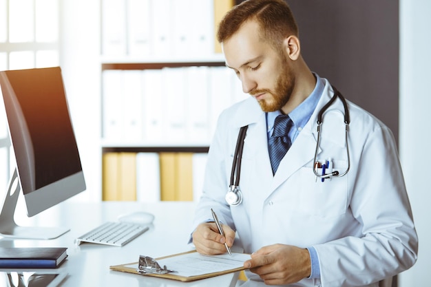 Friendly red-bearded doctor sitting and writing at clipboard in sunny clinic. Medicine concept.