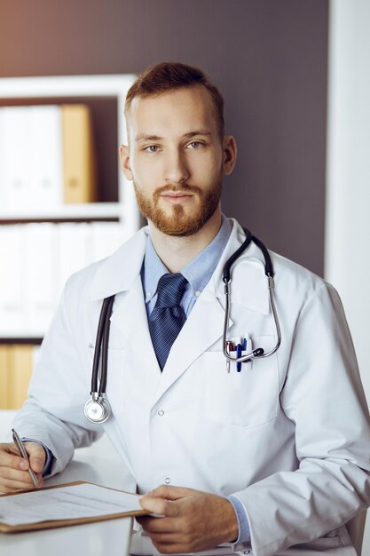 Friendly red-bearded doctor sitting and writing at clipboard in sunny clinic. Medicine concept.