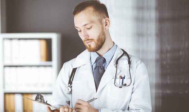 Friendly red-bearded doctor standing and writing with clipboard in clinic at his working place. Perfect medical service in hospital. Medicine concept.