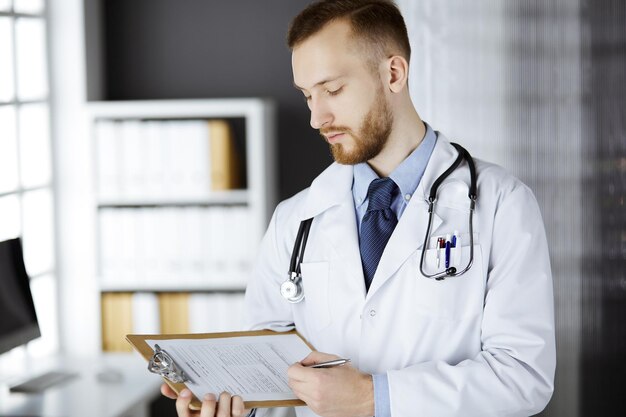 Friendly red-bearded doctor standing and writing with clipboard in clinic at his working place. Perfect medical service in hospital. Medicine concept.