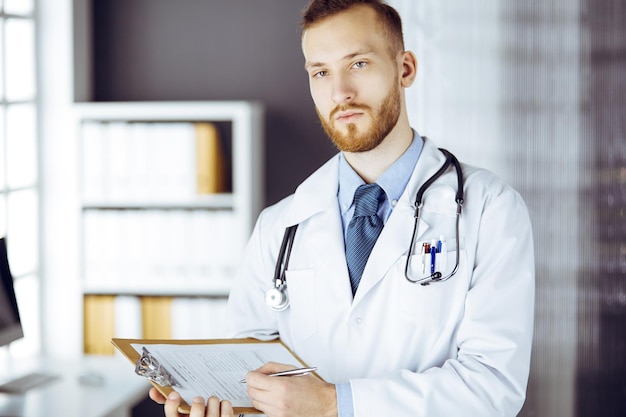 Photo friendly red-bearded doctor standing and writing with clipboard in clinic at his working place. perfect medical service in hospital. medicine concept.