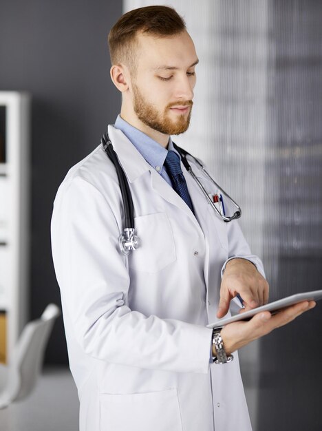 Friendly red-bearded doctor using tablet computer in clinic. Perfect medical service in hospital and medicine concept.