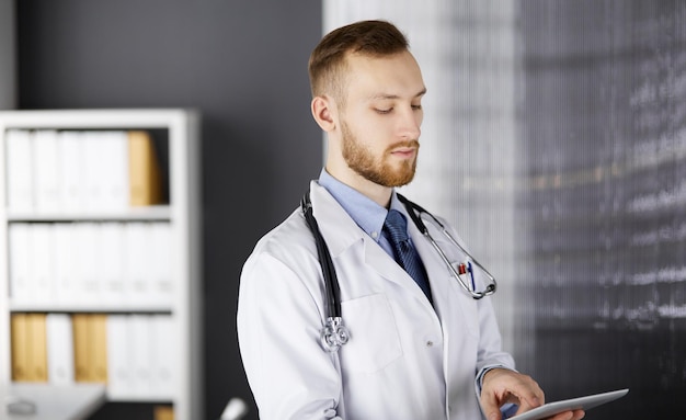 Фото Дружелюбный врач с красной бородой, использующий планшетный компьютер в клинике. идеальное медицинское обслуживание в больнице и концепция медицины.