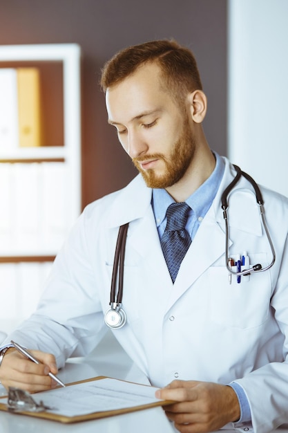 Friendly redbearded doctor sitting and writing at clipboard in sunny clinic Medicine concept