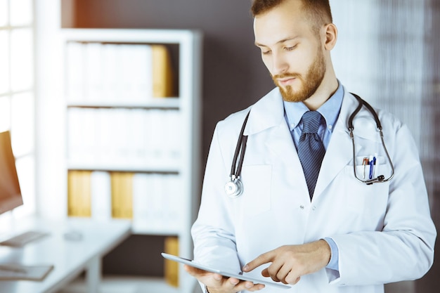 Amichevole medico con la barba rossa che utilizza un computer tablet nella clinica soleggiata nel suo posto di lavoro concetto di medicina