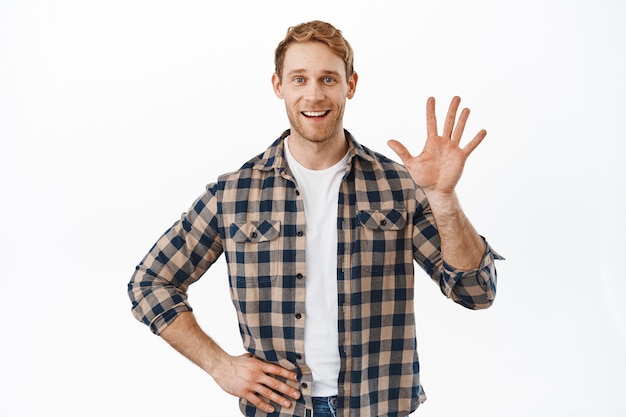 Friendly redhead guy waving hand at you and smiling happy, greeting people, welcome someone, standing against white wall