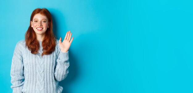 Foto amichevole ragazza adolescente rossa che dice ciao agitando la mano nel gesto di ciao e sorridendo in piedi contro il bl