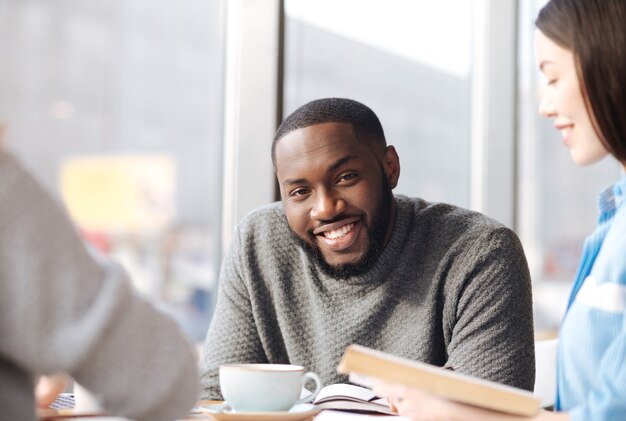 Sorriso amichevole. giovane uomo barbuto bello che sorride e che guarda il suo libro della tenuta dell'amica alla caffetteria.