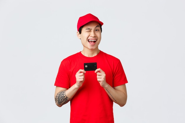 빨간 모자와 티셔츠 유니폼을 입은 친근한 미소 짓는 아시아인 배달원, 쾌활한 윙크, 신용 카드 사용 권장, 바이러스 발생 시 비접촉식 구매. 택배는 코로나 바이러스에 온라인으로 조언 주문