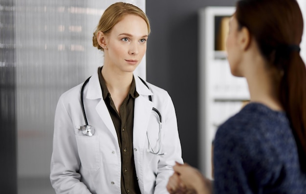 Friendly smiling female doctor and patient woman discussing current health examination while sitting in clinic. Medicine and healthcare concept.