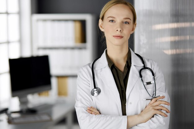 Friendly smiling female doctor standing in clinic. Portrait of friendly physician woman. Perfect medical service in hospital