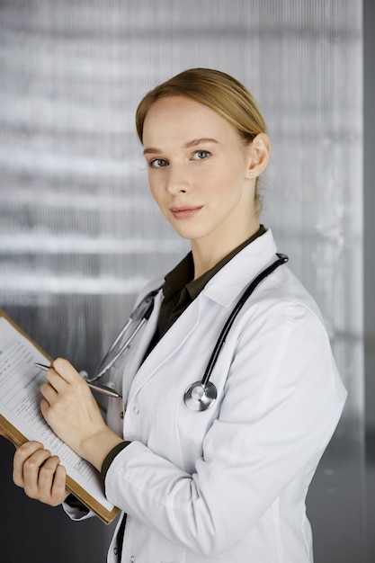 Friendly smiling female doctor using clipboard in clinic. Portrait of friendly physician woman at work. Medical service in hospital. Medicine concept.