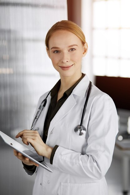 햇볕이 잘 드는 클리닉에서 태블릿 컴퓨터를 사용하는 친절한 웃는 여성 의사. 직장에서 의사 여자입니다. 병원에서 완벽한 의료 서비스. 의학 개념입니다.