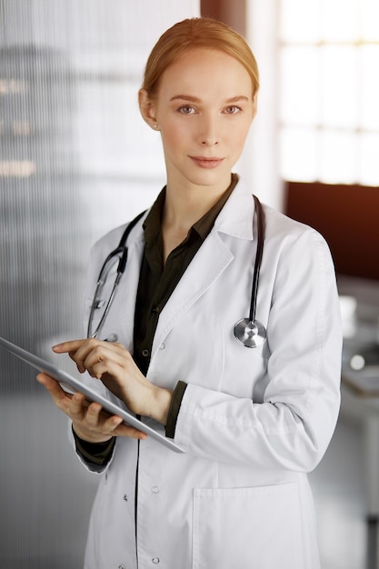 Friendly smiling female doctor using tablet computer in sunny clinic. Physician woman at work. Perfect medical service in hospital. Medicine concept.