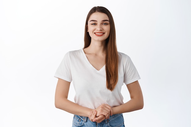 Friendly smiling female student standing in assertive polite pose, ready to assist, talking to client, standing in t-shirt against white wall