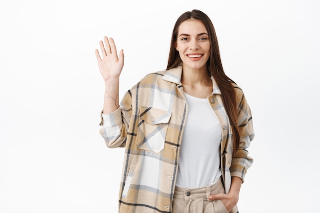 Amichevole donna elegante agitando la mano e salutandoti, salutandoti, dì ehi come va, incontrando persone e dando il benvenuto, in piedi sul muro bianco