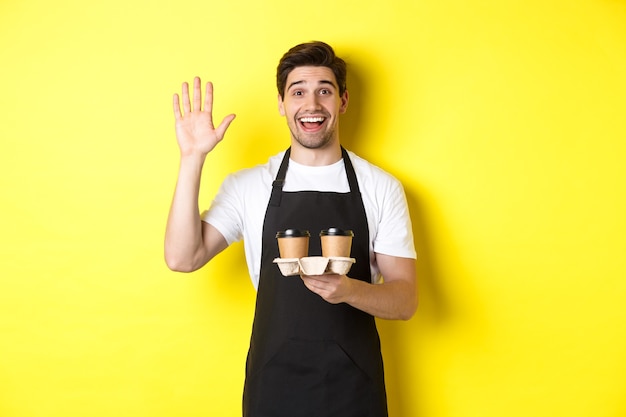 Friendly waiter in cafe waving hand at customer, holding takeaway coffee oder, standing against yellow wall in black apron.