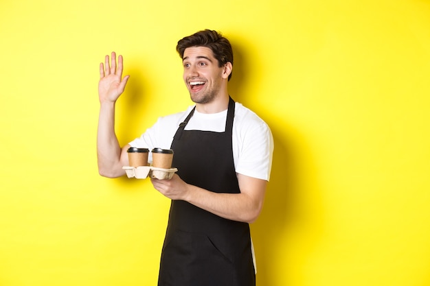 Friendly waiter in cafe waving hand at customer, holding takeaway coffee oder, standing against yellow wall in black apron.