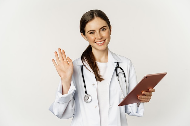 친절한 여성 의사 의사가 손을 흔들며 디지털 태블릿을 들고 행복한 스탠드를 들고 인사합니다...