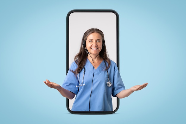 Friendly woman doctor in uniform and headphones spreading arms to sides in big smartphone on blue studio background