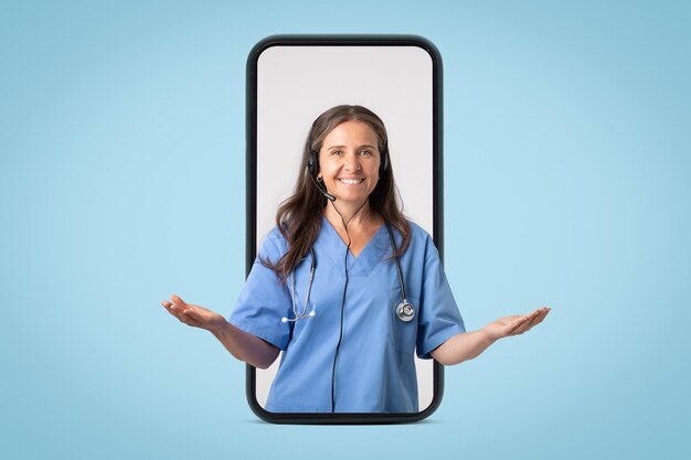 Friendly woman doctor in uniform and headphones spreading arms to sides in big smartphone on blue