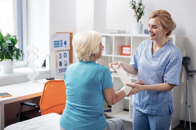 Friendly worker. Positive friendly nurse standing near her patient while bandaging her hand