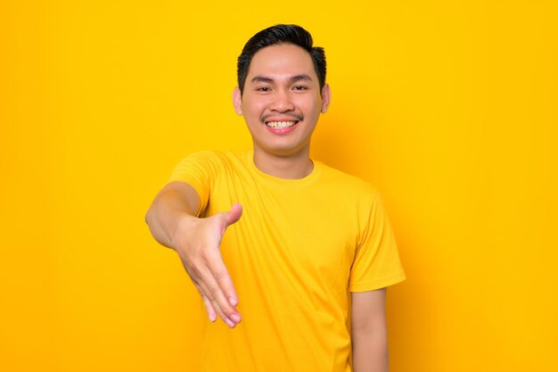 Friendly young asian man in casual tshirt outstretched hand for greeting isolated on yellow background people lifestyle concept