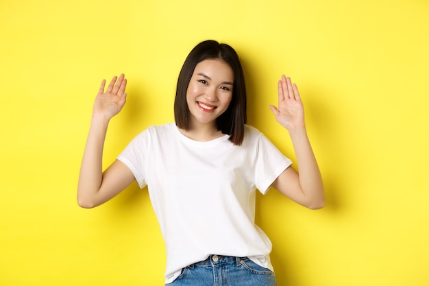 Friendly young asian woman saying hello, raising empty hands up and smiling, greeting you, standing over yellow.