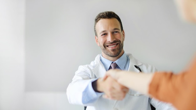 Friendly young man doctor smiling and shaking female patient hand cheerful male therapist greeting