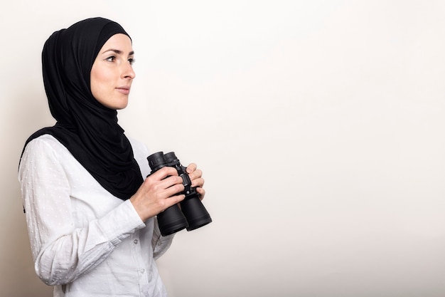 흰 셔츠와 히잡에 친절한 젊은 무슬림 여성이 쌍안경을 보유하고 있습니다.