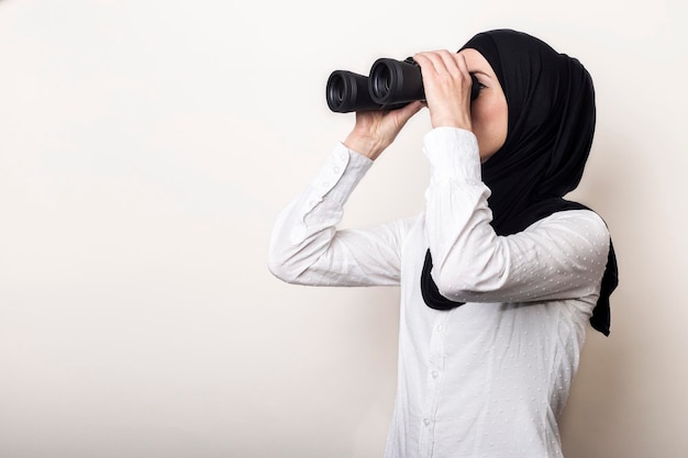 Friendly young Muslim woman in a white shirt and hijab turned to the side looks through binoculars Banner