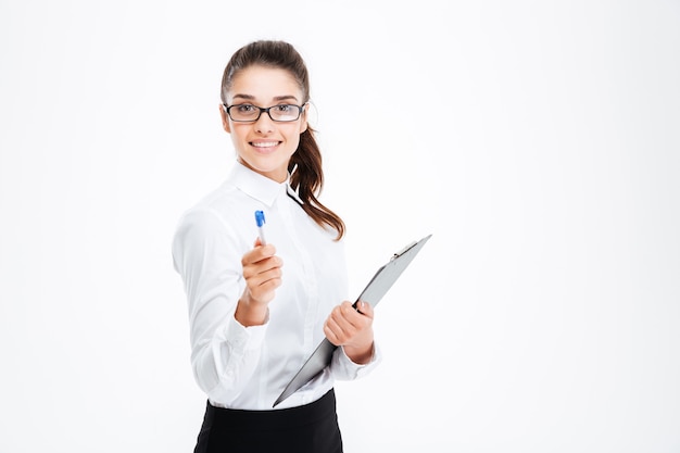 Friendly young smiling businesswoman with clipboard pointing pen at front isolated on white wall