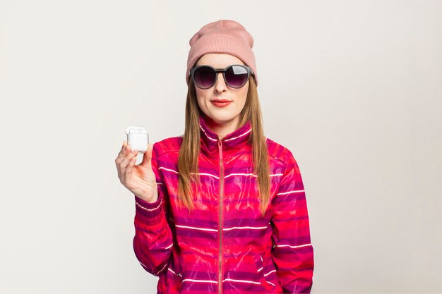 Friendly Young woman in glasses, hat and pink sports jacket with a smiley face holds wireless headphones.