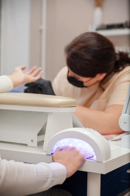 Friendly young woman working in beauty salon as manicure master covering nails of mature female client with gel polish