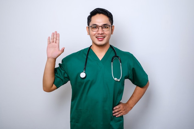 Friendlylooking smiling asian male doctor physician in scrubs waving hand to say hi hello greeting patient