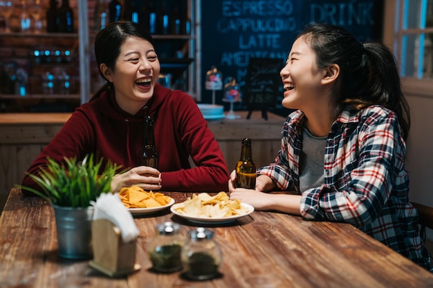 Friends in cafe pub at night drinking beer talking having fun. two asian women sisters enjoy single last day in bar eating chips laughing chatting. happy college girls friends celebrating graduation.