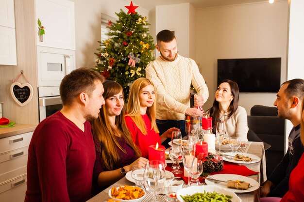 Friends celebrating Christmas or New Year eve sitting in stylish dining room around served table with a lot of dishes, lighted candles. Man standing with bottle of champagne pouring it into glasses