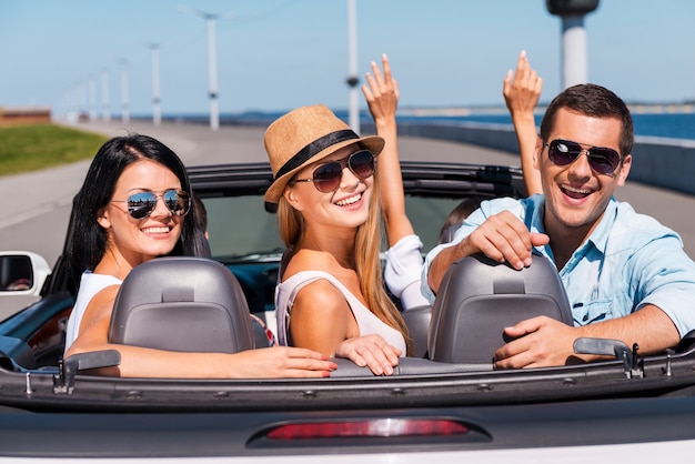 Friends in convertible. Group of young happy people enjoying road trip in their convertible while three of them looking over shoulder and smiling