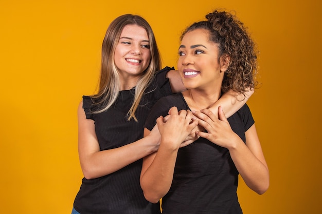 Friends of different ethnicities holding each other on yellow background, diversity concept. An afro woman and a blonde hugging each other smiling looking at the camera