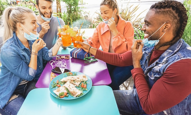 コロナウイルスから保護されるフェイスマスクをつけた夏の日の外のレストランバーでカクテルを飲み、軽食のタパスを食べている友人