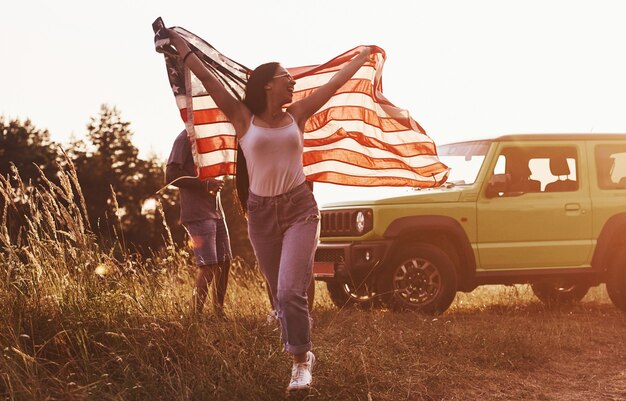 Друзья хорошо проводят выходные на свежем воздухе возле своей зеленой машины с флагом США.
