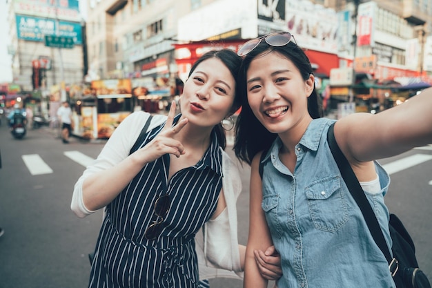 야외에서 즐거운 시간을 보내는 친구들. 자화상을 만드는 얼룩말 십자가 옆에 선글라스를 낀 두 명의 아시아 소녀 여행자. 도시의 도시 도로 대만 타이페이에서 셀카를 찍는 귀여운 여성 관광객