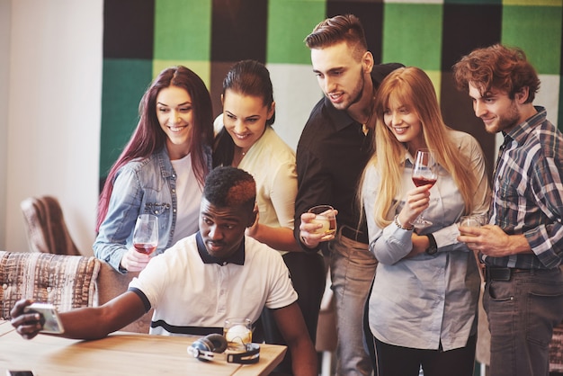 Friends having fun at restaurant. Three boys and three girls making selfie and laughing. On foreground boy holding smart phone. All wear casual clothes