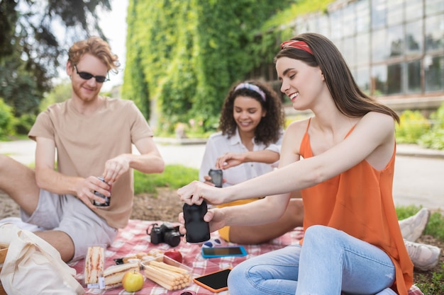 자연의 친구. 소풍 때 공원에서 담요에 앉아 두 여자와 남자 오프닝 음료를 의사 소통하는 즐거운 젊은