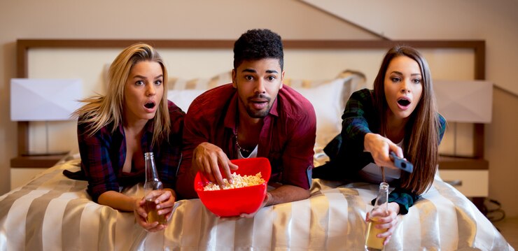 Photo friends lying on bed, having snacks and drinks watching horror movie