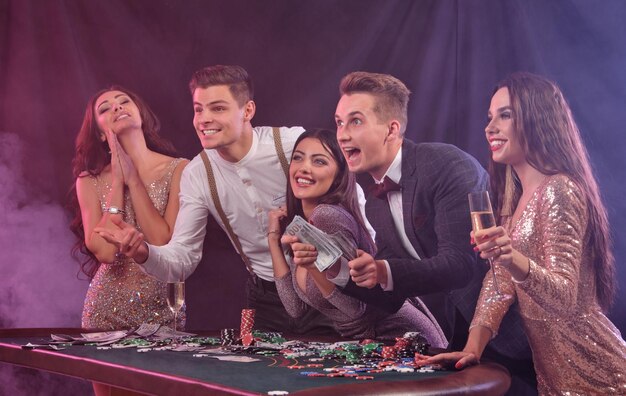 Friends playing poker at casino at table with stacks of chips money cards champagne on it Celebrating their win smiling Black smoke background Gambling entertainment Side view Closeup