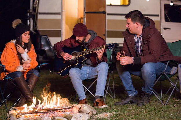 Friends relaxing and playing on guitar around camp fire with their retro camper van in the background. Light bulbs.