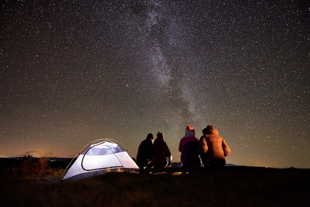 Friends resting beside camp, campfire under night starry sky