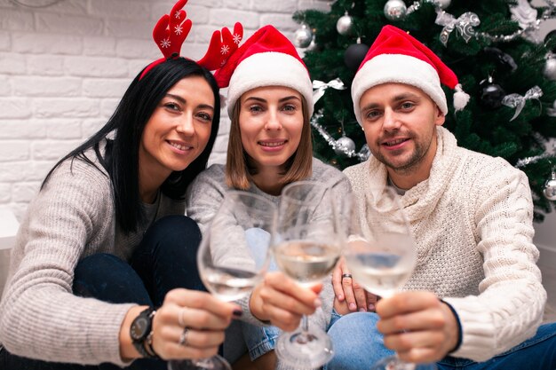 friends in Santa hats sitting near Christmas tree drinking wine